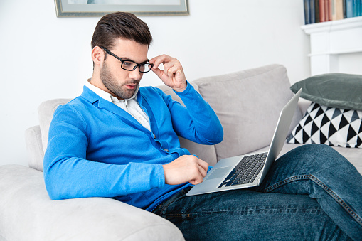 Young male client waiting psychology therapy sitting on sofa touching eyeglasses browsing laptop reading article