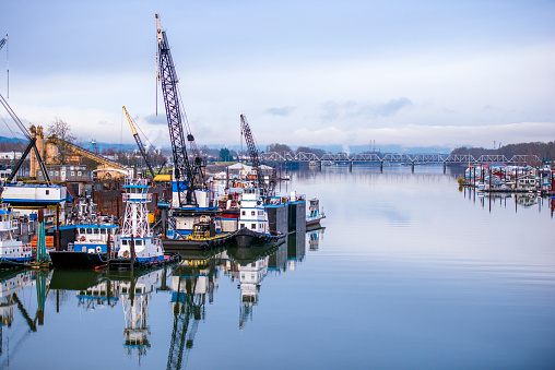 Industrial pier with floating docks on the Columbia River, moored river tugs, cranes for unloading and loading barges on one side and floating houses on the other side of the river and a drawbridge