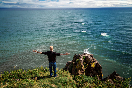 Redhead man in a black sweater stands with arms outstretched near a cliff and looks into the distance to the Pacific Ocean. Freedom, travel, hiking and adventure concept.