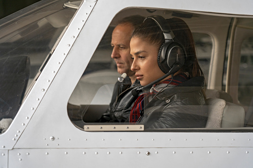 Portrait of attractive young woman trainee pilot with headset preparing to fly. She is sitting next to istructor and looking at dashboard of the private plane.