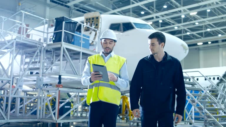 Aircraft Maintenance Mechanic and Chief Engineer Uses Digital Tablet Computer Walk through Big Airplane Development Facility. They Analyze, Inspect, Develop and Design Airplanes