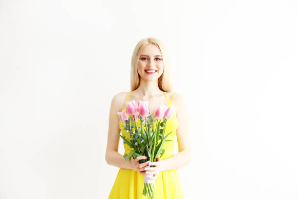 Studio portrait of gorgeous young blonde woman with long straight hair wearing bright yellow dress and holding bouquet of many tender pink tulips. Gray isolated background, copy space, close up.