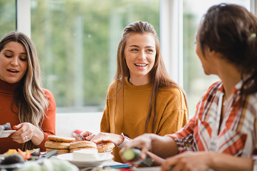 young female adult talking to her friend while having a healthy lunch. They are sitting inside of a conservatory, enjoying themselves.