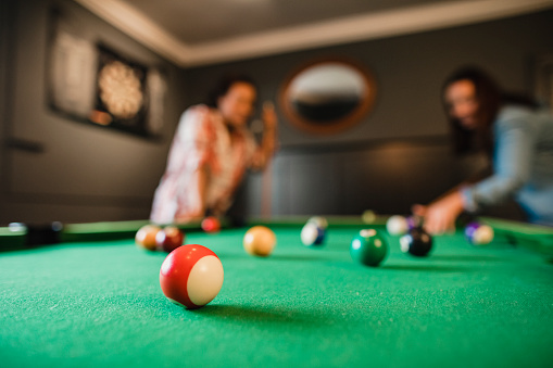Focus on foreground of a red striped pool ball. In the background there are two mid adult friends playing a game of pool in a games room in a house.