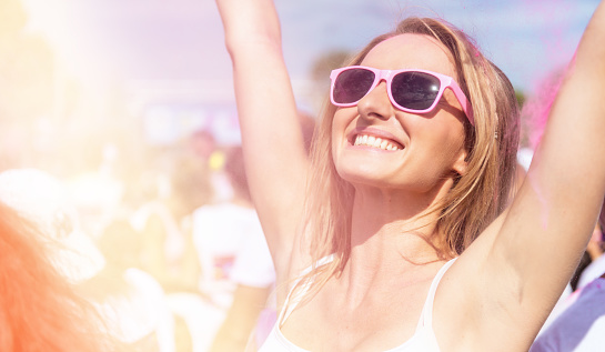 Woman loving the color festival, laughing and screaming with arms raised in crowd of people outdoors. Young candid girl wearing pink sunglasses and enjoying the event.