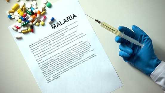 Malaria word typed on paper, doctor hand with syringe pills and tablets on table