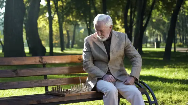Mature man preparing chessboard for game sitting on bench in park, retirement