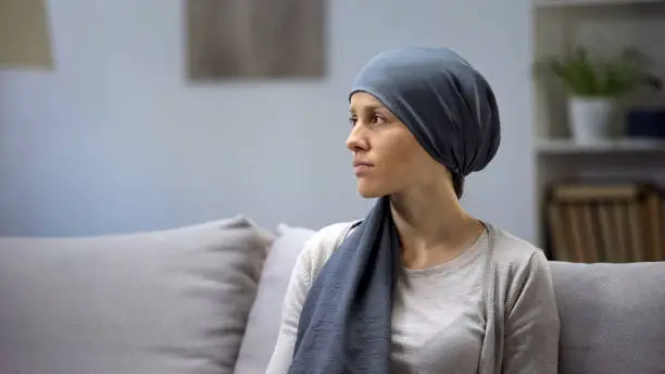 Upset young lady in headscarf lonely sitting on sofa in oncologic hospital