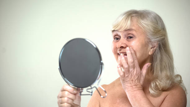 istock Optimistic elderly lady applying anti-aging cream looking in mirror, feminine 1143239427