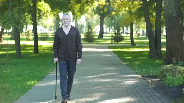 Senior man with walking stick strolling along park breathing fresh air, enjoying
