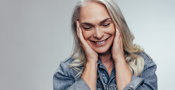 Senior woman with hands on her face and smiling against grey background. Mature woman loving her skin.
