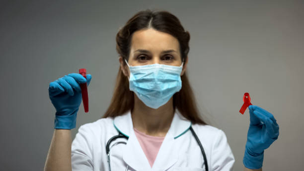 istock Lab technician in protective mask showing red ribbon and blood samples, AIDS 1143240180