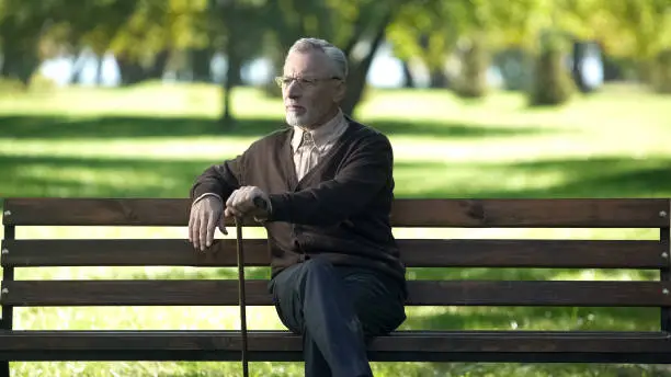 Aged man with walking stick resting on bench in park, enjoying spring nature