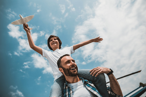 Low angle portrait of laughing bearded man and positive kid having fun with plastic toy under blue sky with white fluffy clouds
