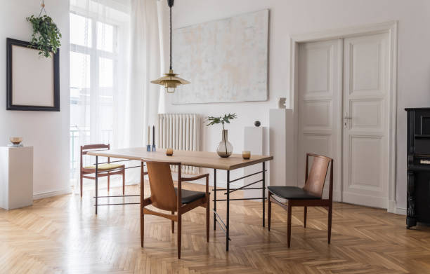 istock Stylish and modern dining room interior with design sharing table, chairs, gold pendant lamp, abstract paintings and elegant accessories. Tropical leafs in vase. Eclectic decor. Brown wooden parquet. 1143252151