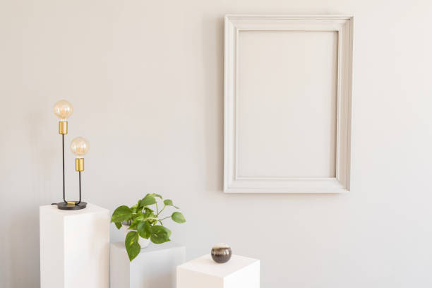 istock Scandi and design home decor with mock up gray poster frame, white and grey pedestals with stylish accessories, lamp and plants in pot. Stylish and minimalistic room interior. Template. Blank. 1143256055