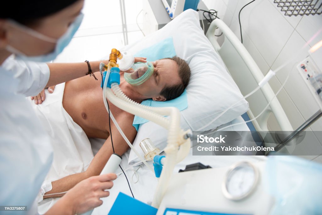 Male patient receiving mechanical ventilation in intensive care ward Top view portrait of middle aged man lying in hospital bed while doctor putting oxygen mask on him Air Duct Stock Photo
