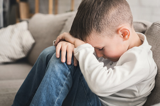 Crying child boy hugging his knees, sitting on sofa, closeup