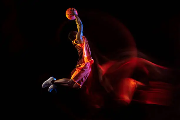 Catching the sun. African-american young basketball player of red team in action and neon lights over dark studio background. Concept of sport, movement, energy and dynamic, healthy lifestyle.