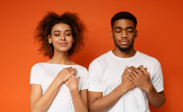 Kind woman and man keep both palms on chest Grateful african-american couple keep both palms on chest with closed eyes, express gratitude and good feeling, orange background black woman praying stock pictures, royalty-free photos & images