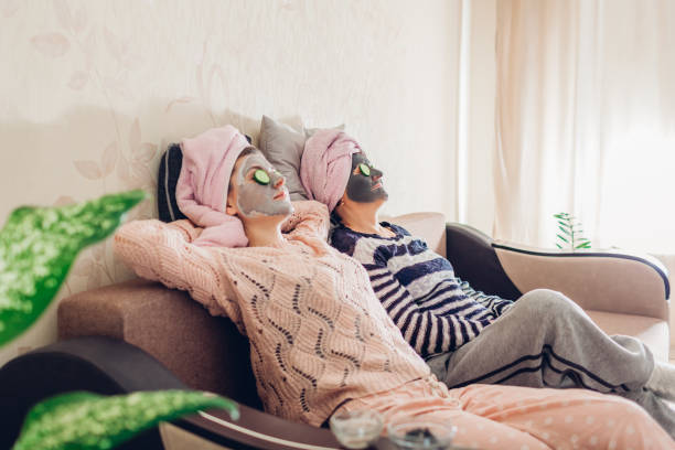 madre y su hija adulta relajándose con máscaras faciales y pepinos aplicados. las mujeres escalofriante y divertirse en casa - facial mask spa treatment cucumber human face fotografías e imágenes de stock