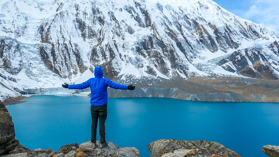Young man standing in front of Tilicho Lake with his arms wide spread. Blue and calm surface of the lake, mountains covered in the shadow, sunlight in the back. Overcast. Harsh landscape. Thankfulness