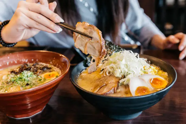 Woman hand pinching noodle in Ramen Pork Bone Soup (Tonkotsu Ramen) with Chashu Pork, Scallion, Sprout, Corn, Dried Seaweed and boiled egg served in black bowl.