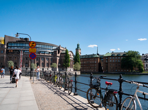 Tourists and local residents walking past bicycles which are chained to railings, with the Swedish parliament building (Riksdaghuset) and Storkyrken (Stockholm cathedral) prominent. Various buildings in Gamla stan, Stockholm’s old town,  are in the background to the right.