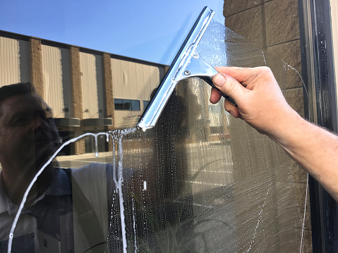 A close up of a window cleaner's arm holding a squeegee to clean a store window at an office building or store front. The window washer's reflection is in the window. The squeegee is wiping the water and soap suds away to reveal a clean window.