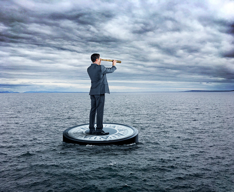 A businessman stands on a large compass as he looks through a spyglass while navigating through a sea that is beginning to turn rough.