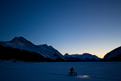 A man goes for a nighttime winter fat bike ride on a frozen lake in the Rocky Mountains of Canada. Fat bikes are mountain bikes with oversized wheels and tires for riding on the snow. He has lights on his bicycle and his head to light up the trail.