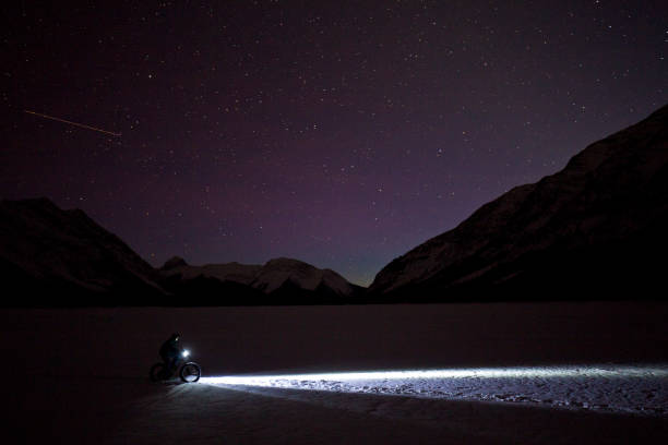 Fat Biking Under the Stars A man goes for a nighttime winter fat bike ride under the stars in the sky on a frozen lake in the Rocky Mountains of Canada. Fat bikes are mountain bikes with oversized wheels and tires for riding on the snow. He has lights on his bicycle to light up the trail. bicycle light photos stock pictures, royalty-free photos & images