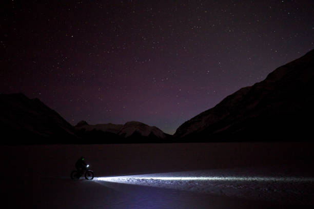 Fat Biking Under the Stars A man goes for a nighttime winter fat bike ride under the stars in the sky on a frozen lake in the Rocky Mountains of Canada. Fat bikes are mountain bikes with oversized wheels and tires for riding on the snow. He has lights on his bicycle to light up the trail. bicycle light photos stock pictures, royalty-free photos & images