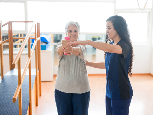 istock Senior woman lifting weights with both arms next to therapist 1143400904