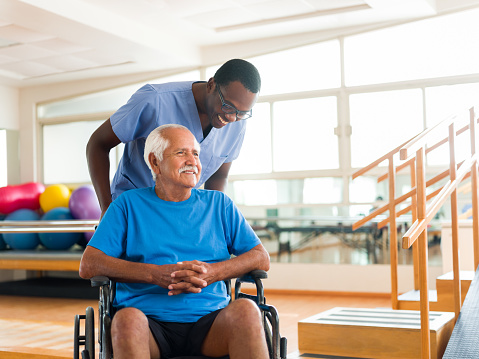 A black male nurse standign behind a senior male patient on a wheelchair and smiling.