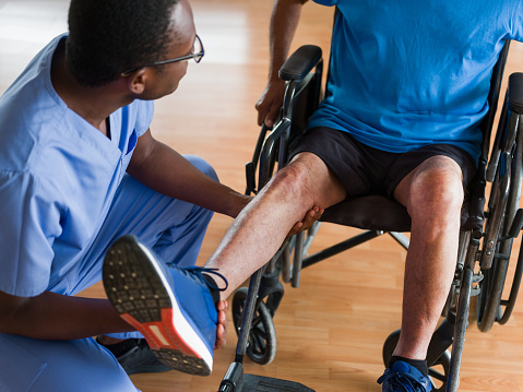 A black male nurse crouching in front of a senior patient sitting on a wheelchair and checking his leg.