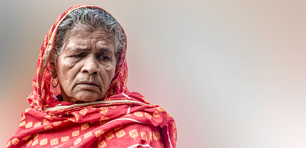 Portrait of an Indian Origin Elderly Women, farmer by profession, wearing traditional Indian dress of red color with veil and thinking deep in sorrow because farming is a very tough job in India in the present scenario. She is in her 70s and still has to work everyday without fail and is hoping for the farm loan waivers. This portrait is kinda very typical face or may say a Poster Face of almost every elderly women of rural India. Might also be taken as an elderly woman suffering from Dementia or Memory loss because of the blurred background.