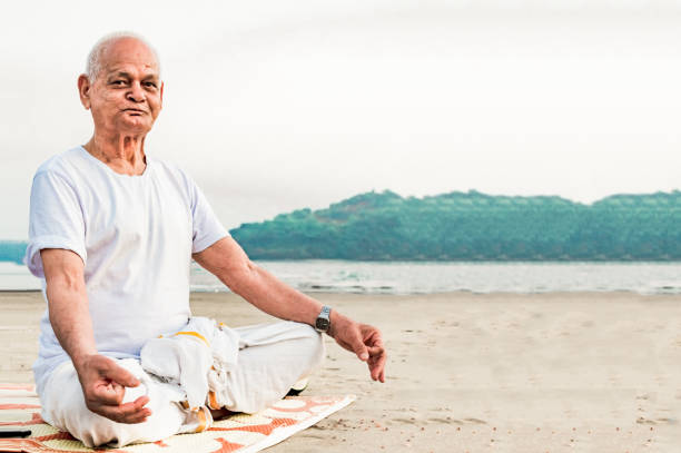 Healthy Old Indian Man, Super Senior Citizen performing Yoga, sitting in Lotus Pose and doing meditation on beach. He is wearing dhoti, has pretty wrinkles on his face and is happily posing for camera Yoga Guru or Yoga Teacher of Indian origin in his early 90s is performing yoga by sitting in Lotus Pose of meditation on a natural beach of South India. He is superannuated and is living a very healthy lifestyle without any ailments or disease, as he leads a spiritual and yoga lifestyle. kerala photos stock pictures, royalty-free photos & images