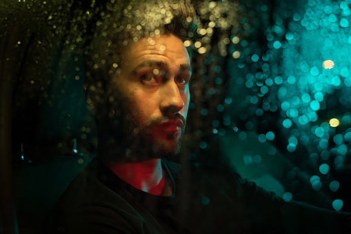 Young man driving at night, under the rain, illuminated with colorful street lights.