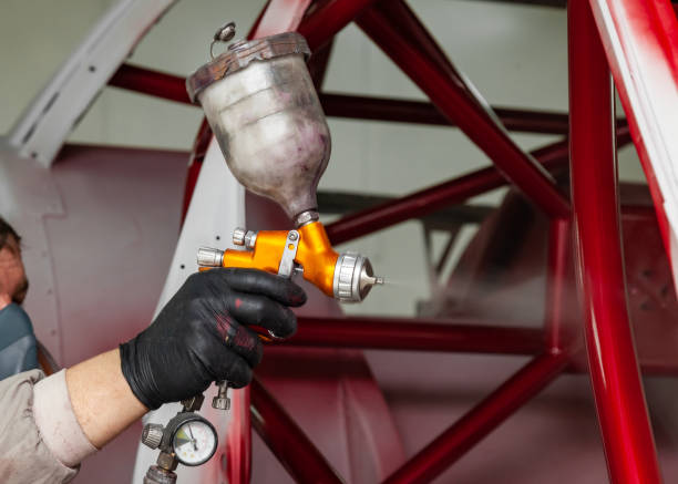 un uomo padrone in tuta protettiva e una maschera tiene in mano una bomboletta spray e spruzza vernice rossa sul telaio della carrozzeria dopo un incidente durante una riparazione in un'officina di restauro del veicolo - priming paint foto e immagini stock