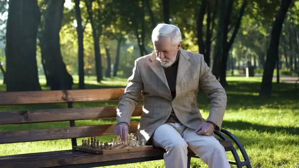 Old man sitting on bench in park alone and playing chess, retirement benefits