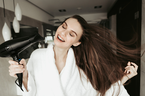 Concept of haircare and enjoyment after shower. Close up portrait of cheerful beautiful woman closing eyes while drying hair with blowdryer at home bathroom interior