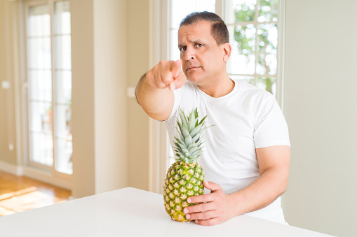 Middle age man eating fresh tropical pineapple at home pointing with finger to the camera and to you, hand sign, positive and confident gesture from the front