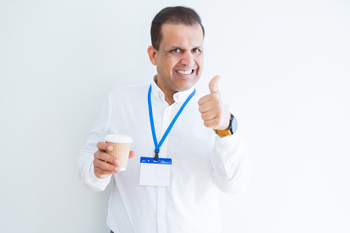 Middle age business man wearing ID card and drinking coffeeover white background happy with big smile doing ok sign, thumb up with fingers, excellent sign