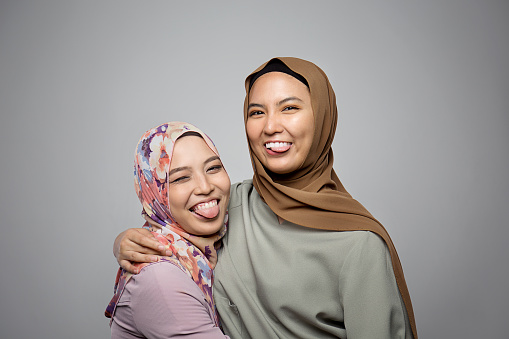 Two Young Muslim girls wearing hijab sticking out tongue and smiling in front of camera isolated on grey