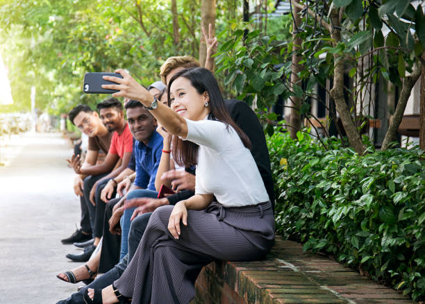 istock Young Asian friends sitting outdoors on the low brick wall and taking a selfie 1143479385