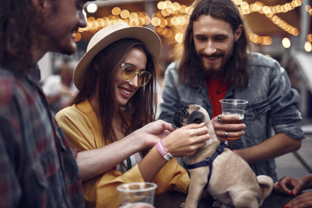 Young people petting little pug while standing at the table Our four-legged friend. Portrait of stylish hipster friends looking at cute dog and smiling. Bearded guy holding beer hipster dog stock pictures, royalty-free photos & images