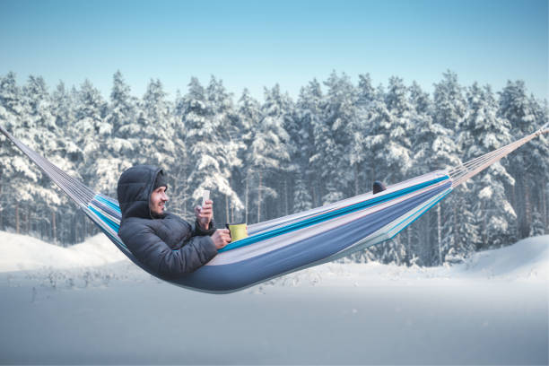 un utilisateur de smartphone se trouve dans un hamac dans une forêt enneigée hivernale. mug avec une boisson chaude dans sa main - hamac telephone homme photos et images de collection