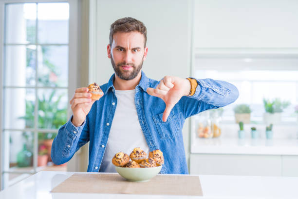 istock Handsome man eating chocolate chips muffin with angry face, negative sign showing dislike with thumbs down, rejection concept 1143482843