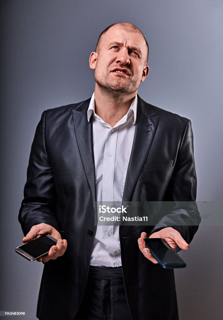 Unhappy stressed tired business man holding two mobile phones in hands and looking up in office suit on grey studio background. Closeup Unhappy stressed tired business man holding two mobile phones in hands and looking up in office suit on grey studio background. Closeup portrait Adult Stock Photo